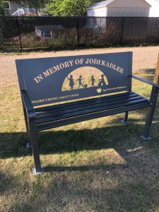 Image of one of Age-Friendly Rosetown's commemorative benches, in a grassy area. It says In Memory of Jodi Kadler.