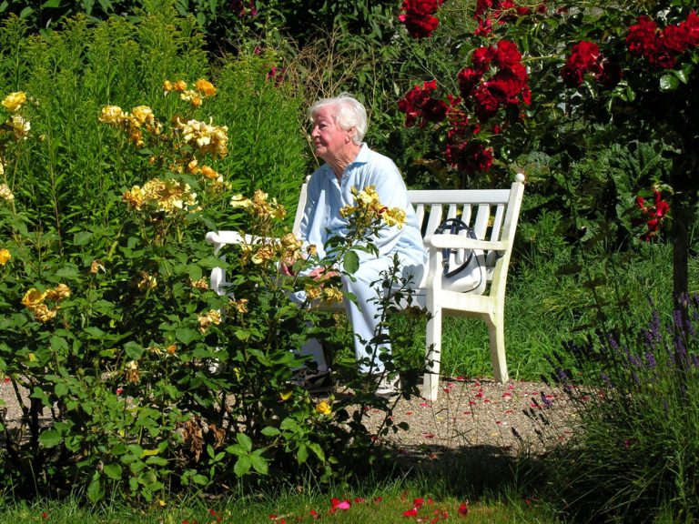 Older woman sitting on bench in flower garden.