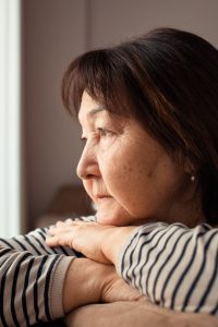 Image of pensive older Asian or Indigenous woman, seated with arms folded across the back of the couch, her chin on her hands as she looks out the window.