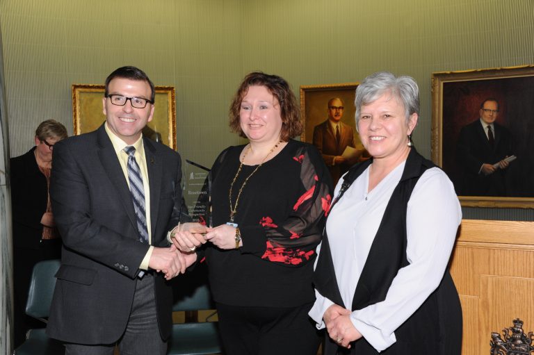 Photo of two women from Age-Friendly Rosetown receiving their recognition award from the government minister. Portraits of past premiers are on the walls behind them.