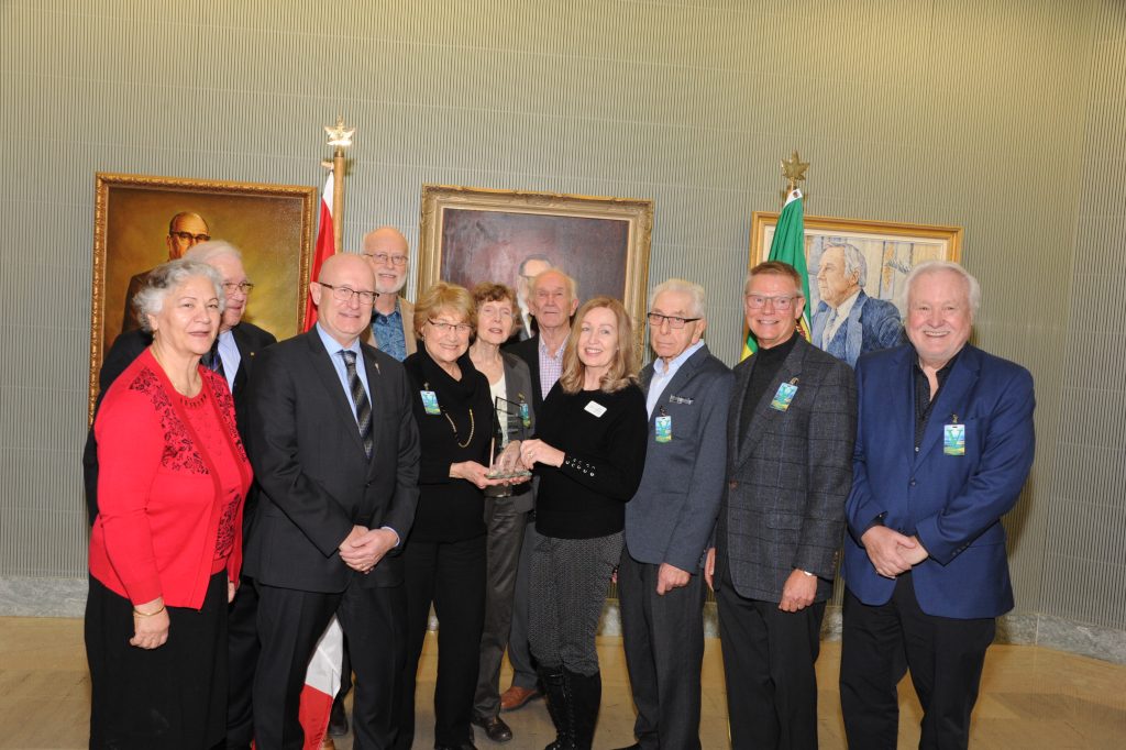 Photo of Age-Friendly Saskatoon representatives and SSM representatives as Age-Friendly Saskatoon accepted their recognition award from the Saskatchewan Government. Photos of past premiers are on the wall behind them.
